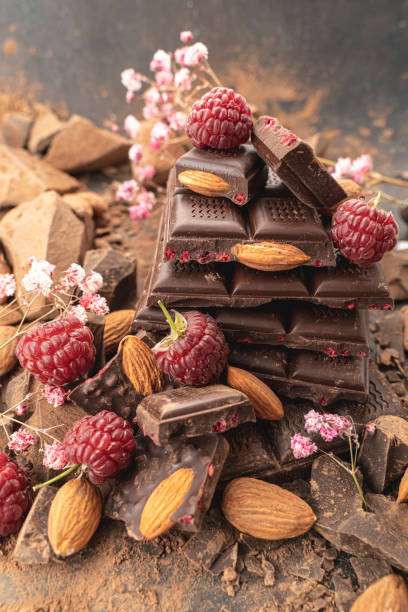 istock Cooking chocolate bars with almonds, raspberries, handmade sweets. Composition of bars and pieces dark chocolate, raspberry, flowers on black background top view. Confectionery craft on table 1348974572