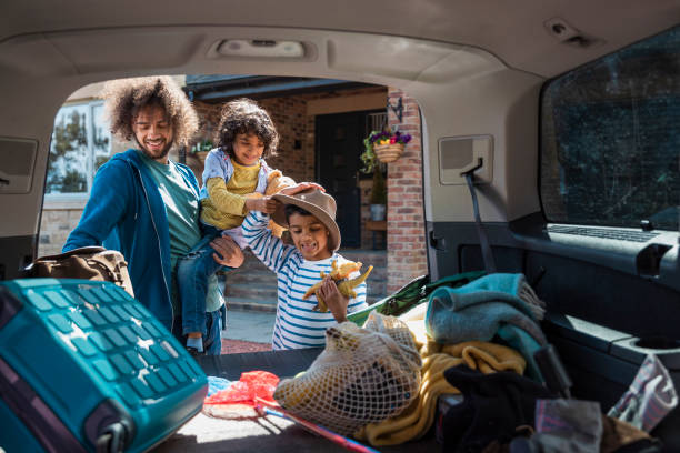 Starting our Holiday Staycation Medium shot with camera placed inside of a car showing a man and his son unpacking a car boot, getting their bags and toys when they arrive at the lodge they're staying on during a staycation in the Northeast of England together. The young boy is trying on his fathers hat, having fun and the man is holding one of the boys. northern europe family car stock pictures, royalty-free photos & images