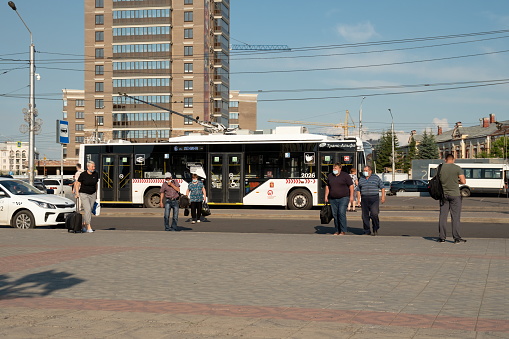 Krasnoyarsk, Krasnoyarsk Region, RF - July 11, 2021: People in medical masks with bags walk from the bus stop from the trolleybus past a taxi against the backdrop of city houses on a sunny summer day.