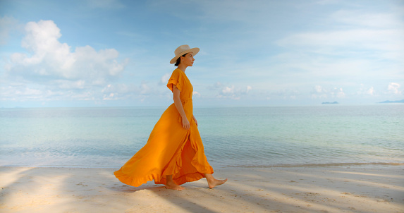 Beautiful girl in yellow long dress and hat walking at the edge of water and looking at the calm sea.