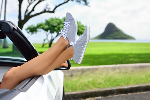 Relaxing vacation car road trip travel with feet up the window. Convertible car holiday concept with female legs in front of Oahu landmark : Mokoli’i in Hawaii, USA.