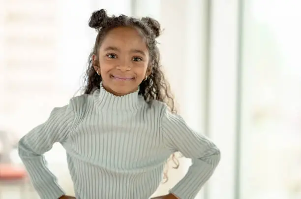 A young girl of African decent stands in front of a bright picture window and casually poses with her hands on her hips.  She is wearing a light blue sweater and has half her hair up in buns as she smiles.