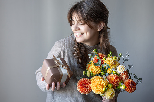 Young brunette woman smiling with a bright bouquet of fresh chrysanthemums and a gift box in her hands, on a gray background.