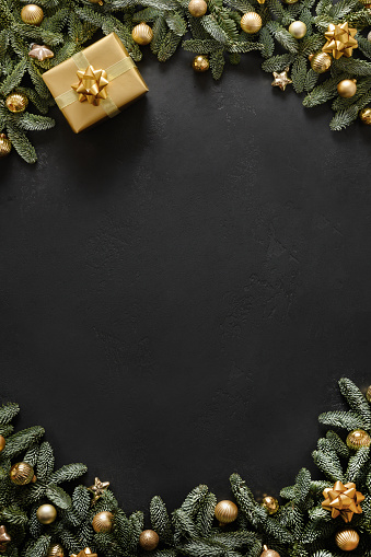 Christmas vertical frame with golden gift, baubles, evergreen branches on black background with copy space. Xmas greeting card. Happy New Year. View from above, flat lay.