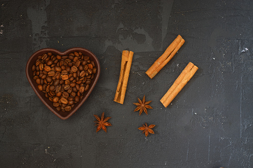 Roasted coffee beans in heart shaped bowl on black stone table. Love coffee concept. Top view flat lay with copy space. With Star anise and cinnamon sticks.