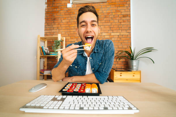 Front view of cheerful handsome young man eating sushi looking or reading information from monitor screen, looking at camera.