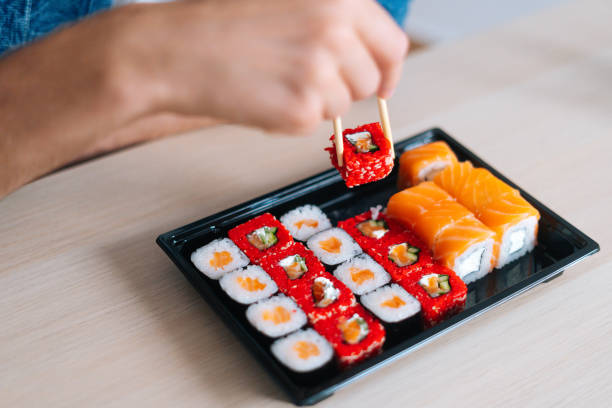 Close-up high-angle view hands of unrecognizable young man holding sushi with chopsticks sitting at desk