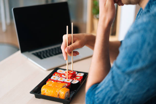 Close-up back view of unrecognizable young man eating sushi rolls with chopsticks sitting at working desk with laptop at home office.