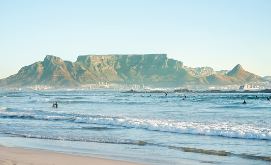Ocean laps beach coastline with people surfing. Table Mountain, Lion's Head & Devil's Peak across the bay. Cape town, South Africa.