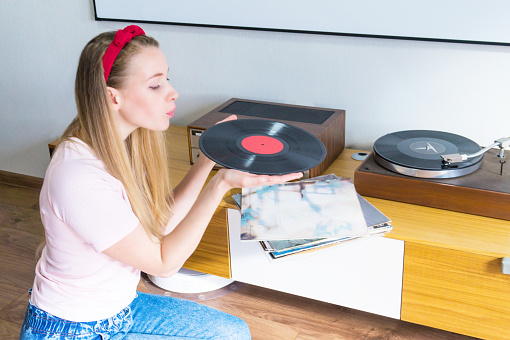 Young woman blowing dust off vinyl record and listening to music on turntable at home.
