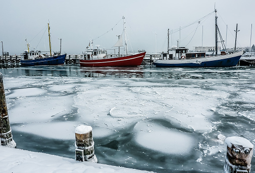 Side view of three fishing boats in the icy water in the harbor of rostock, Germany. Sailing boats moored in the harbor by the buildings against the cloudy sky.