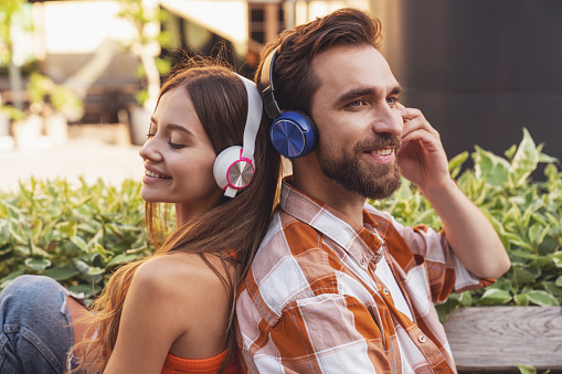 Young smiling couple listening to music on headphones sitting on a park bench back to back.