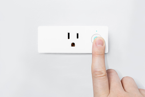 Finger pressing an on-off button with blue light active for safety testing electric system on modern smart plug and wifi outlet. The minimal design of electricity with control switch for saving energy on a wall.