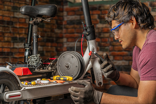 A latin mechanic male with protective goggles sitting and using his cell phone while repair an electric scooter in his workshop with different tools on top of the scooter.