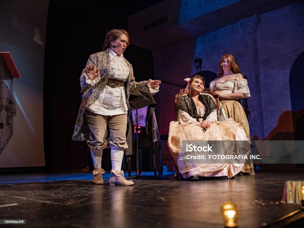 Three Actresses in period costumes on theatre stage Three Actresses in period costumes on theatre stage during opera performance. . Interior of  Theatre stage during performance  at night. Performing Arts Event Stock Photo