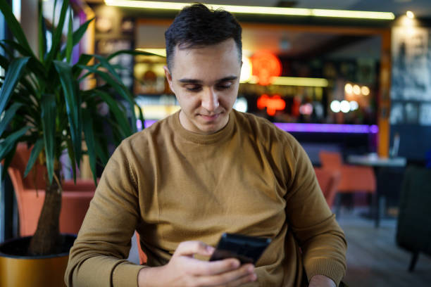 istock Young caucasian man sitting alone at cafe front view of one happy male while using mobile phone for texting or browsing internet or making a phone call copy space real people 1349070915