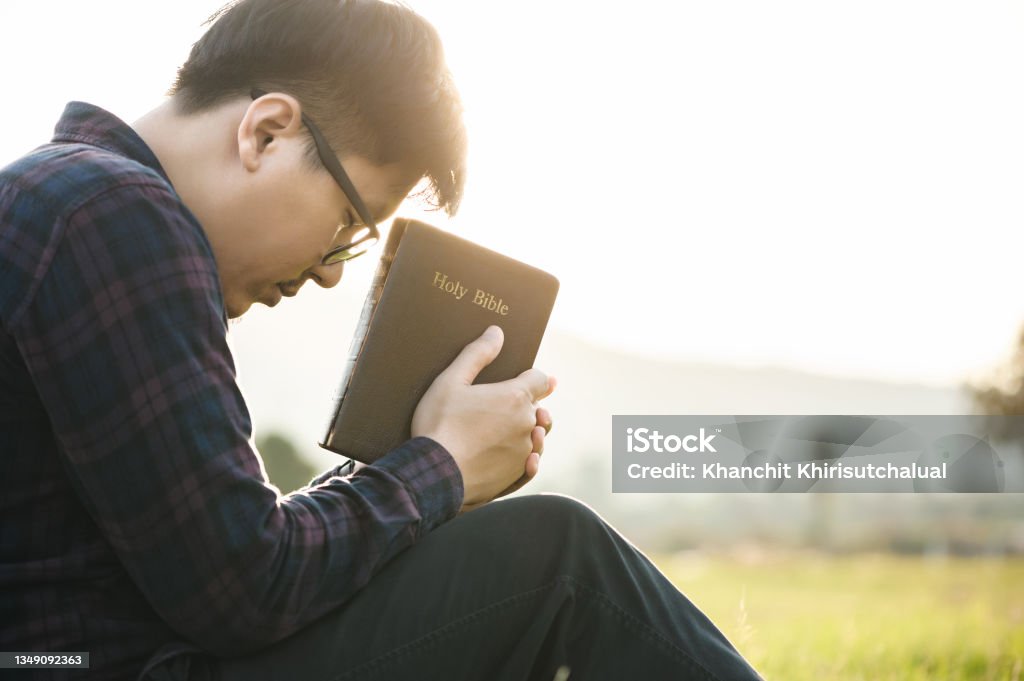 Christian man sit holds the bible in hands to reading the Holy Bible and pray in a field during a beautiful sunset. The concept for faith, spirituality, and religion. Peace, hope.sit Humility Stock Photo