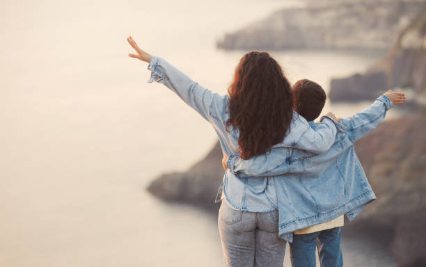 młoda mama i jej nastoletni syn w dżinsowych ubraniach na szczycie góry, spędzają razem czas i przytulają się w wiosenny dzień. hiszpanka i dziecko na wakacjach. szczęśliwa koncepcja relacji rodzinnych. - hiszpanka_& zdjęcia i obrazy z banku zdjęć