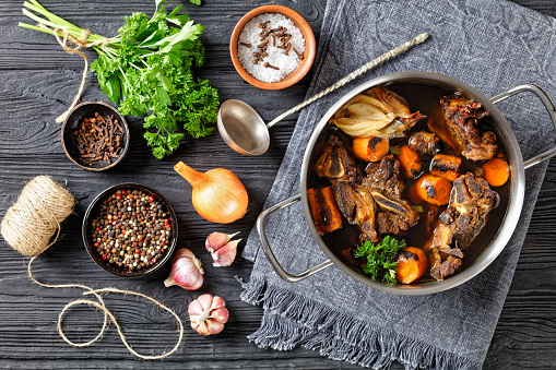 beef broth of beef meat on bones slow cooked with charred vegetables: carrot, onion, garlic, and spices served in a pot on a wooden background with ingredients, top view, flat lay