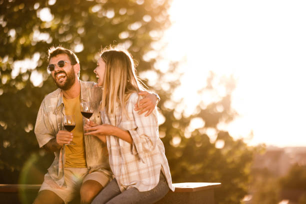 Cheerful couple having fun while drinking wine at sunset. Young cheerful couple having fun while drinking wine and laughing on a roof at sunset. Copy space. couple drinking stock pictures, royalty-free photos & images