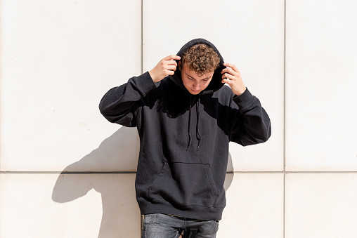 Young man in black sweatshirt putting his hood up on the outside and looking down on a concrete block background