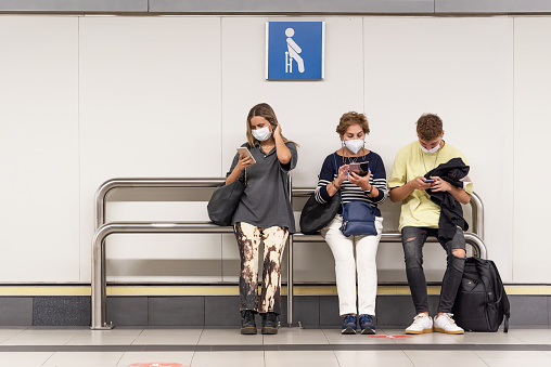 A woman, a man and an older woman sitting at a subway stop looking at their cell phones wearing mask