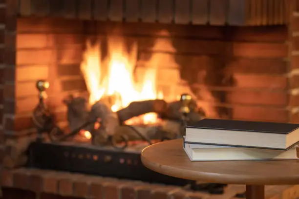 Photo of Books stacked on a small table in front of a fireplace with fire in winter.