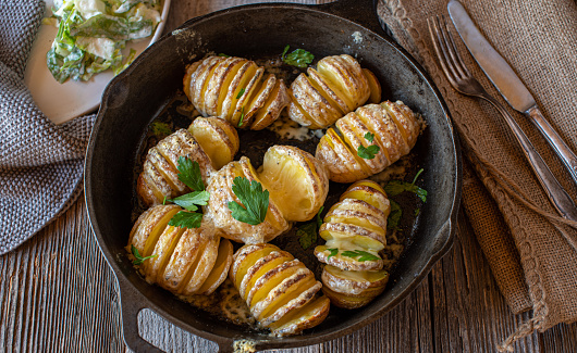 Delicious vegetarian dish or side dish with baked hasselback potatoes with melted butter and cheese. Served in a rustic cast iron pan on wooden table. Overhead view