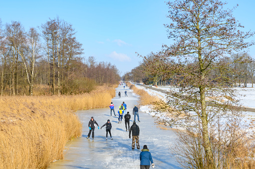 People ice skating on frozen canals and lakes in the Weerribben-Wieden nature reserve in Dwarsgracht village in Overijssel, The Netherlands during a cold winter day. The land and ice is covered in snow under the clear blue sky.