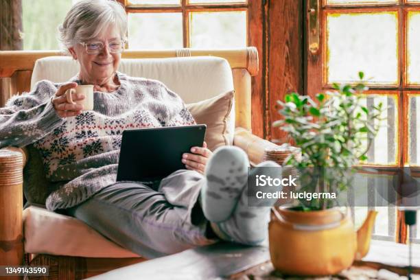 Old Senior Woman Sitting At Home On Armchair Using Digital Tablet Wearing A Warm Sweater And Eyeglasses Comfortable Living Room Wooden Rustic Windows Stock Photo - Download Image Now