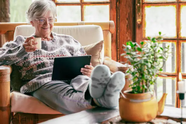 Photo of Old senior woman sitting at home on armchair using digital tablet wearing a warm sweater and eyeglasses. Comfortable living room, wooden rustic windows