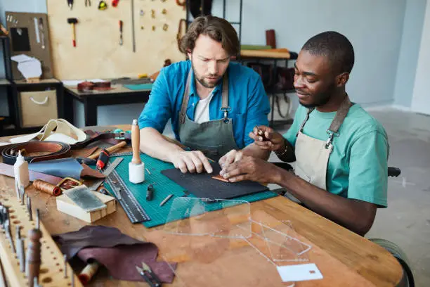 Portrait of male artisan teaching young apprentice using wheelchair in leatherworkers workshop, copy space