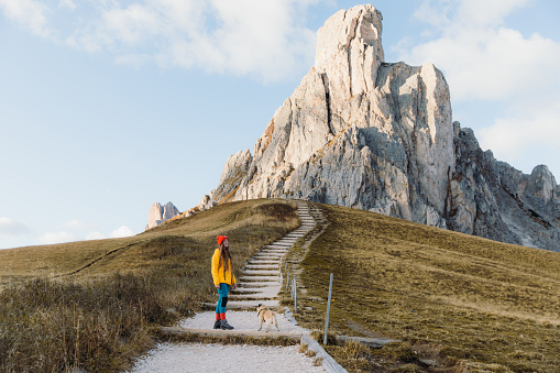 Young happy woman explorer in yellow jacket and red hat with her cute small dog - pug breed hiking through the footpath with scenic view of the beautiful peak during autumn sunset in Dolomites mountains, South Tyrol, Italy