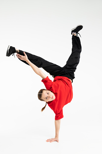 Isolated young Russian girl in red t-shirt hip hop break dancer dancing in studio in white background, performing freeze hand stand of downrock breakdance looking into the camera