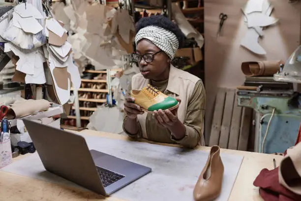 African young shoemaker sitting at the table in front of the laptop and presenting new design shoe online in the workshop