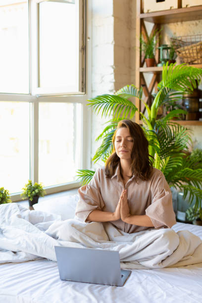 Vertical photo of a adult woman meditating in bed in front of laptop monitor. Yoga practice stock photo