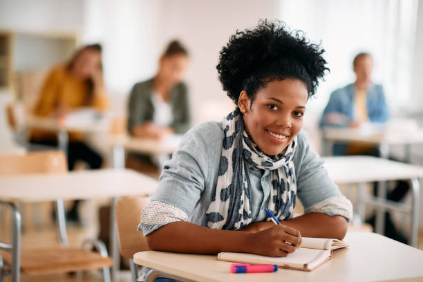 Happy black woman writing an exam while attending a class at the university and looking at camera. Happy African American mid adult student writing while having lecture in the classroom and looking at camera. mature student stock pictures, royalty-free photos & images