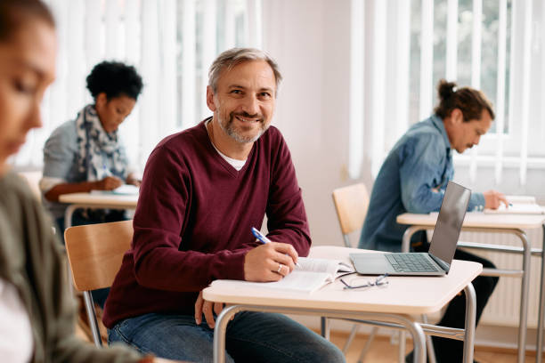 Happy mature student learning in the classroom and looking at camera. Happy mature man attending a lecture and writing notes while looking at camera. mature student stock pictures, royalty-free photos & images