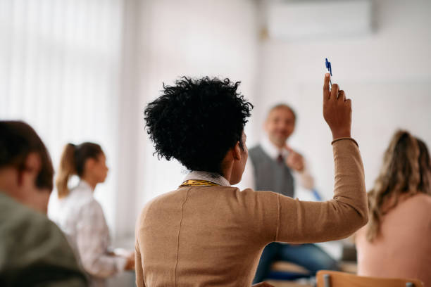 Rear view of black student raising her hand to answer a question during a class at lecture hall. Back view of African American student raising her arm to answer a question during lecture in the classroom. adult student stock pictures, royalty-free photos & images