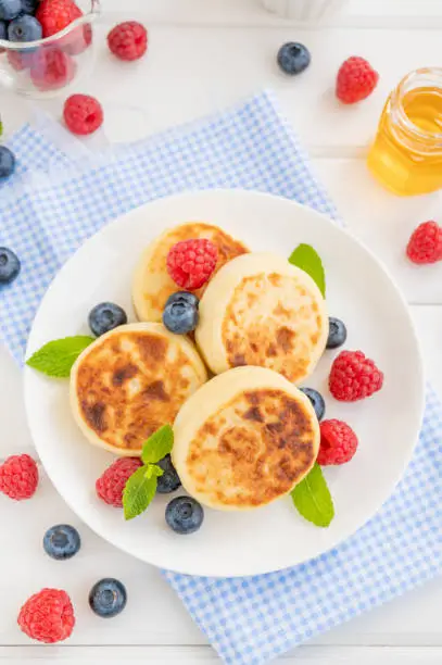 Cottage cheese pancakes with fresh berries, sour cream and honey on a white wooden background. Homemade traditional Ukrainian and Russian syrniki