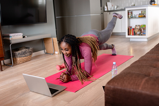 Young African America woman at home doing exercise in front of open laptop, repeating instructions by professional online fitness trainer.