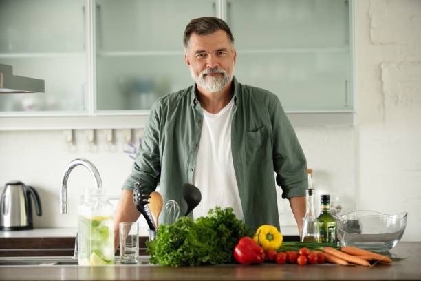 istock Smiling mature man standing in kitchen, looking at camera, making fresh vegetable salad enjoying cooking alone at home 1349177340
