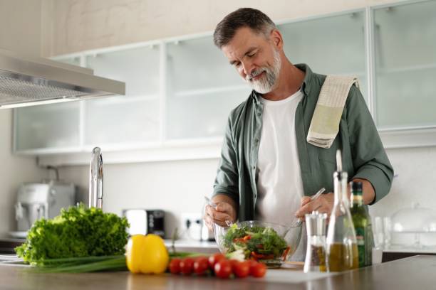 homme mûr heureux mélangeant une salade de légumes frais debout dans la cuisine à la maison. - cooking chef domestic kitchen food photos et images de collection