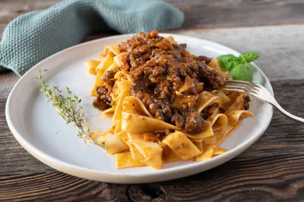 Delicious fresh and homemade cooked italian ragout dish with duck meat, root vegetables, tomatoes and herbs. Served with pasta isolated on a plate on wooden table.