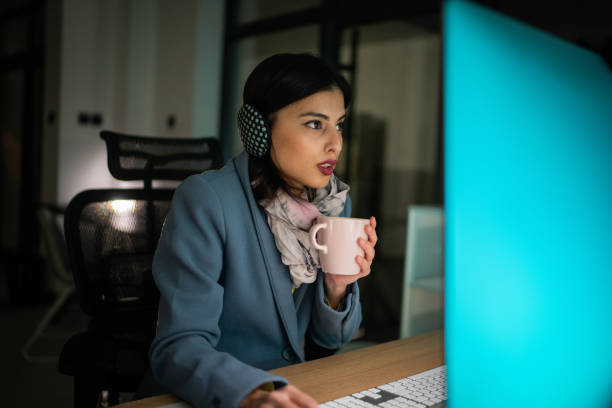 Woman in warm clothing sitting in freezing office and working on computer with hot coffee cup in hands stock photo