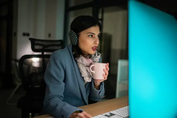 Young woman in warm clothing sitting in freezing office and working on computer with hot coffee cup in hands