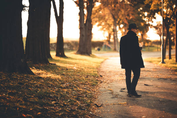 Portrait of a handsome young male model wearing black hat and long black coat, he is walking in autumn public park.