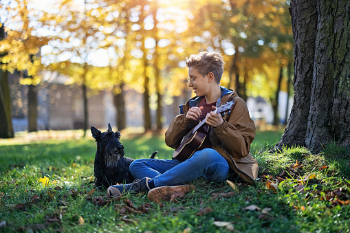 Teenage boy aged 12 playing ukulele for his dog. The boy is sitting on grass, next to a tree in a public park. Beautiful sunny autumn day.\nCanon R5