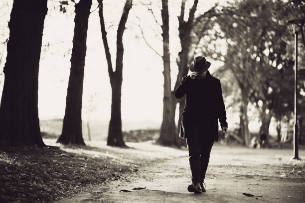 Portrait of a handsome young male model wearing black hat and long black coat, he is walking in autumn public park.