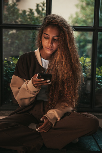 Teenage girl texting on social media sitting by a large window at home